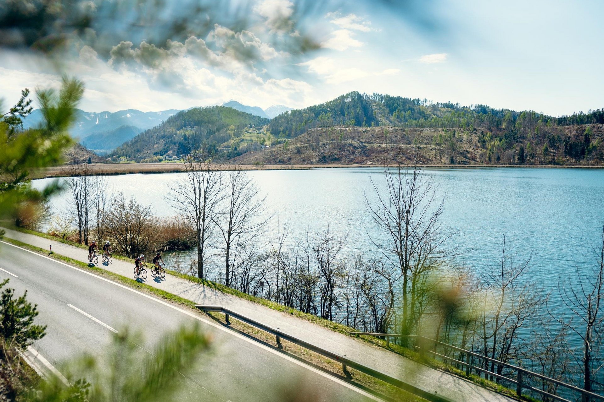 Roadbike Holidays © Suedkaernten Fahrradfahrer auf Straße neben See und Hügeln unter bewölktem Himmel