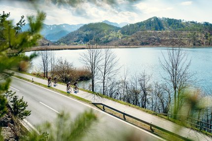Diex - Völkermarkt © Suedkaernten Ciclisti su strada accanto a lago con colline e cielo nuvoloso