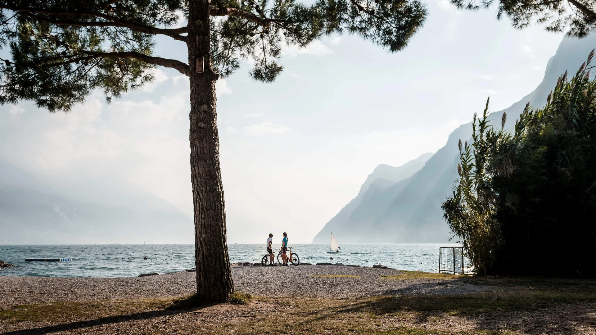 3 Regionen für den Rennradurlaub im Herbst © Alex Moling Zwei Radfahrer am Ufer eines Sees mit Bergen und Segelboot im Hintergrund
