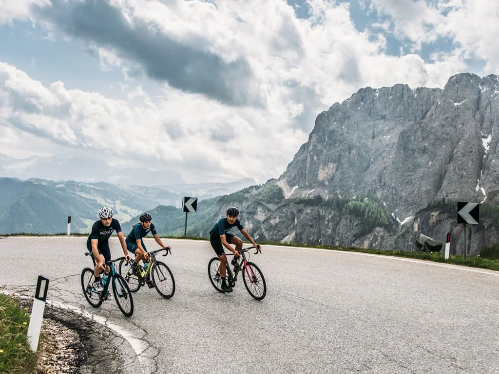 Tre ciclisti su strada di montagna con rocce e cielo nuvoloso