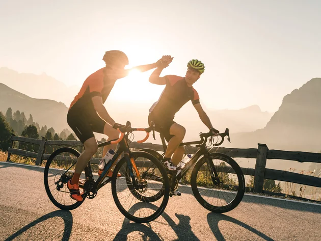 Warum Alta Badia auf jede Roadbike-Bucket-List gehört © Molography Zwei Rennradfahrer geben sich beim Sonnenuntergang High Five auf einer Bergstraße