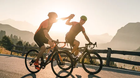 Warum Alta Badia auf jede Roadbike-Bucket-List gehört © Molography Zwei Rennradfahrer geben sich beim Sonnenuntergang High Five auf einer Bergstraße