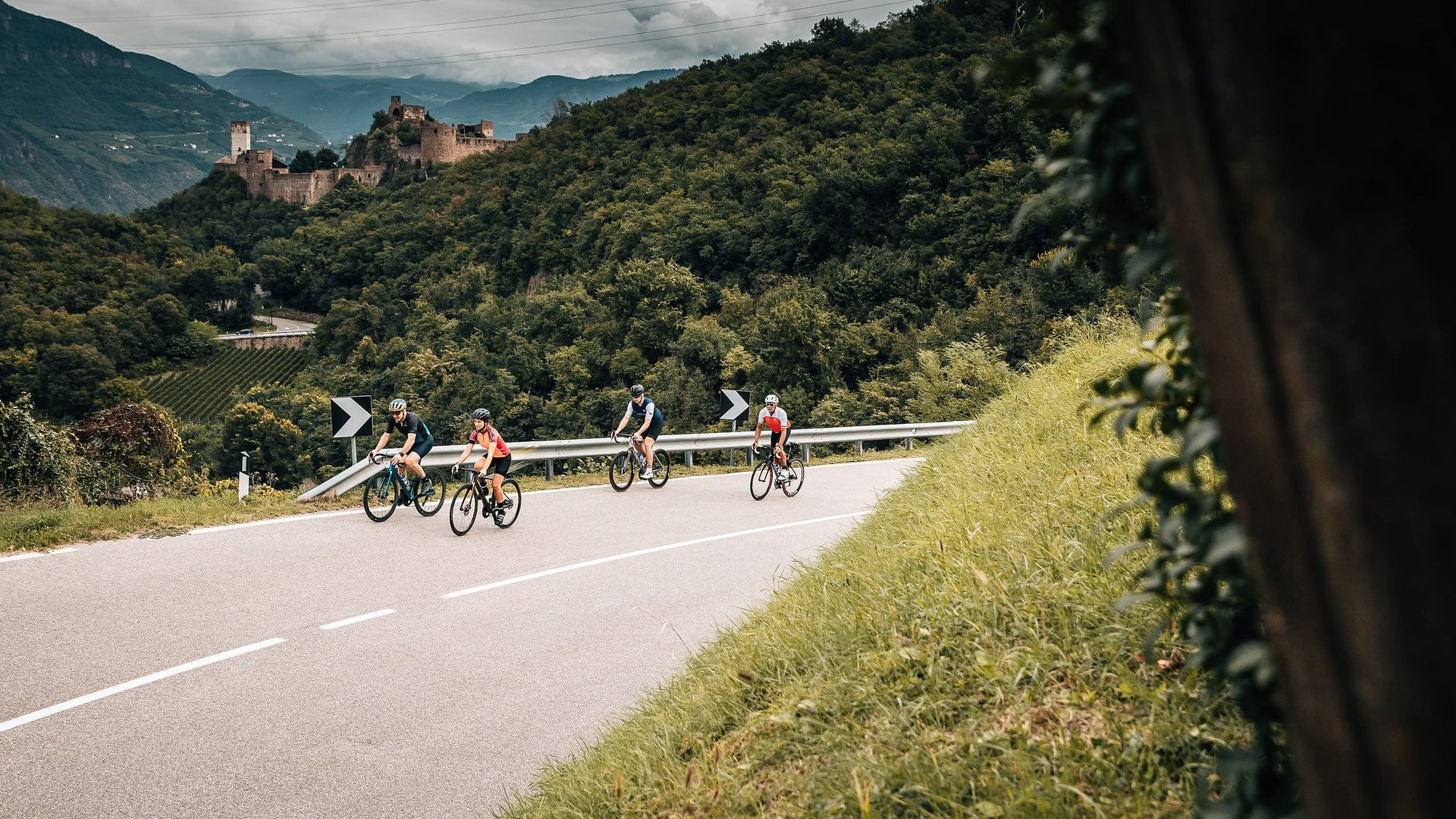 Vier Radfahrer fahren auf kurviger Bergstraße mit bewaldeten Hügeln und Burg