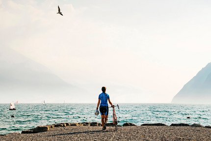 Fahrradfahrer am Seeufer mit Bergen und Segelbooten im Hintergrund