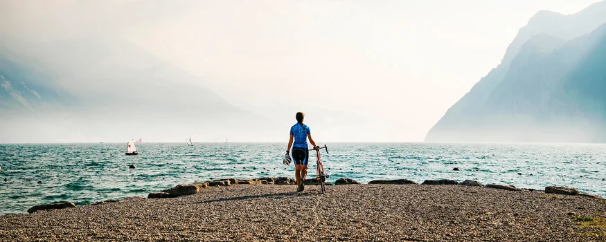 Die schönsten Rennrad-Regionen und Tipps für den Herbst © Alex Moling Fahrradfahrer am Seeufer mit Bergen und Segelbooten im Hintergrund