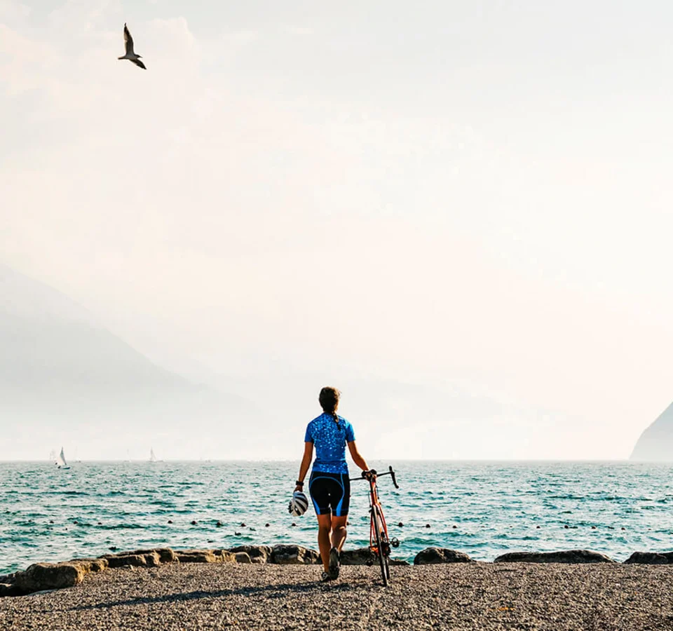 Cyclist standing by lakeside with mountains and sailboats in the background
