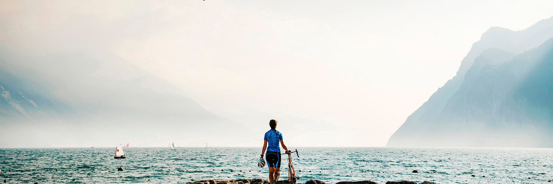 Cyclist standing by lakeside with mountains and sailboats in the background