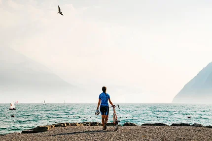 Fahrradfahrer am Seeufer mit Bergen und Segelbooten im Hintergrund