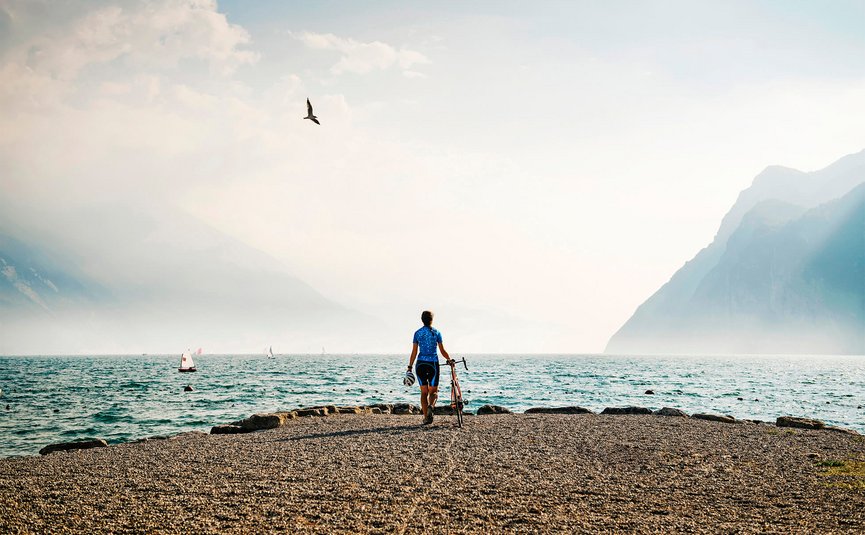 Ciclista al lago con montagne e barche a vela sullo sfondo