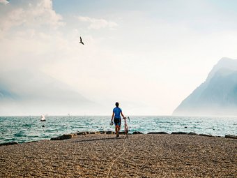 Fahrradfahrer am Seeufer mit Bergen und Segelbooten im Hintergrund