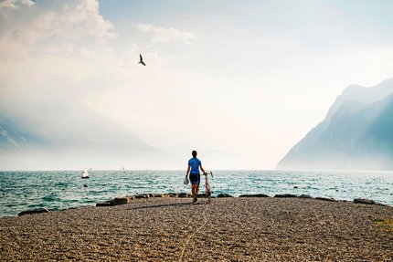 Cyclist standing by lakeside with mountains and sailboats in the background