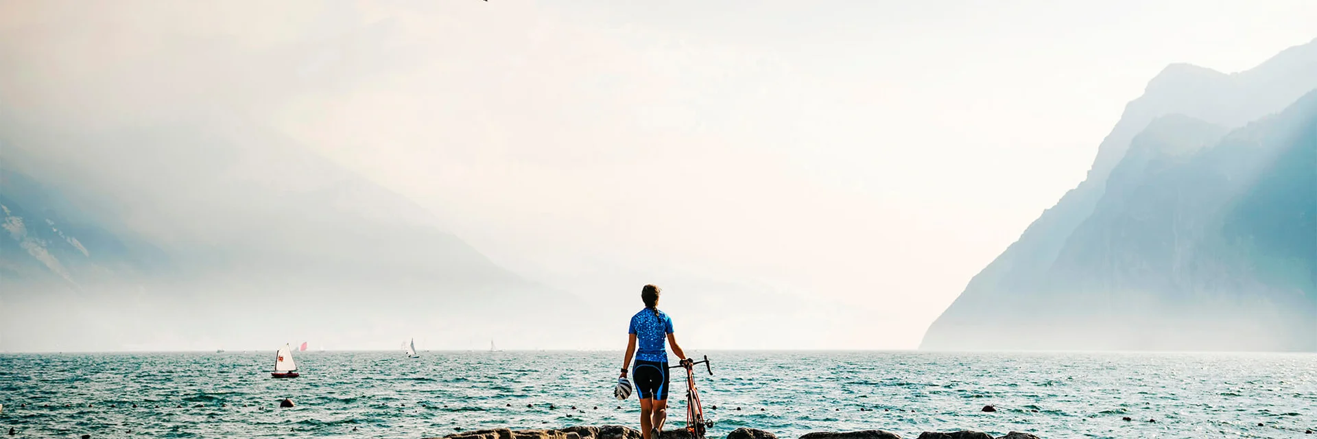 Ciclista al lago con montagne e barche a vela sullo sfondo