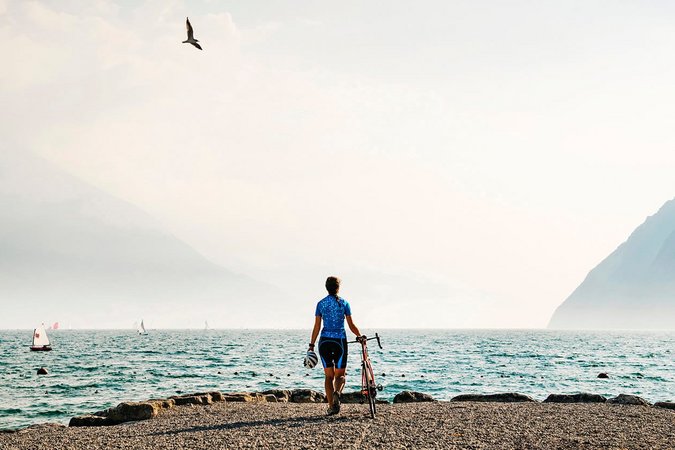 Fahrradfahrer am Seeufer mit Bergen und Segelbooten im Hintergrund