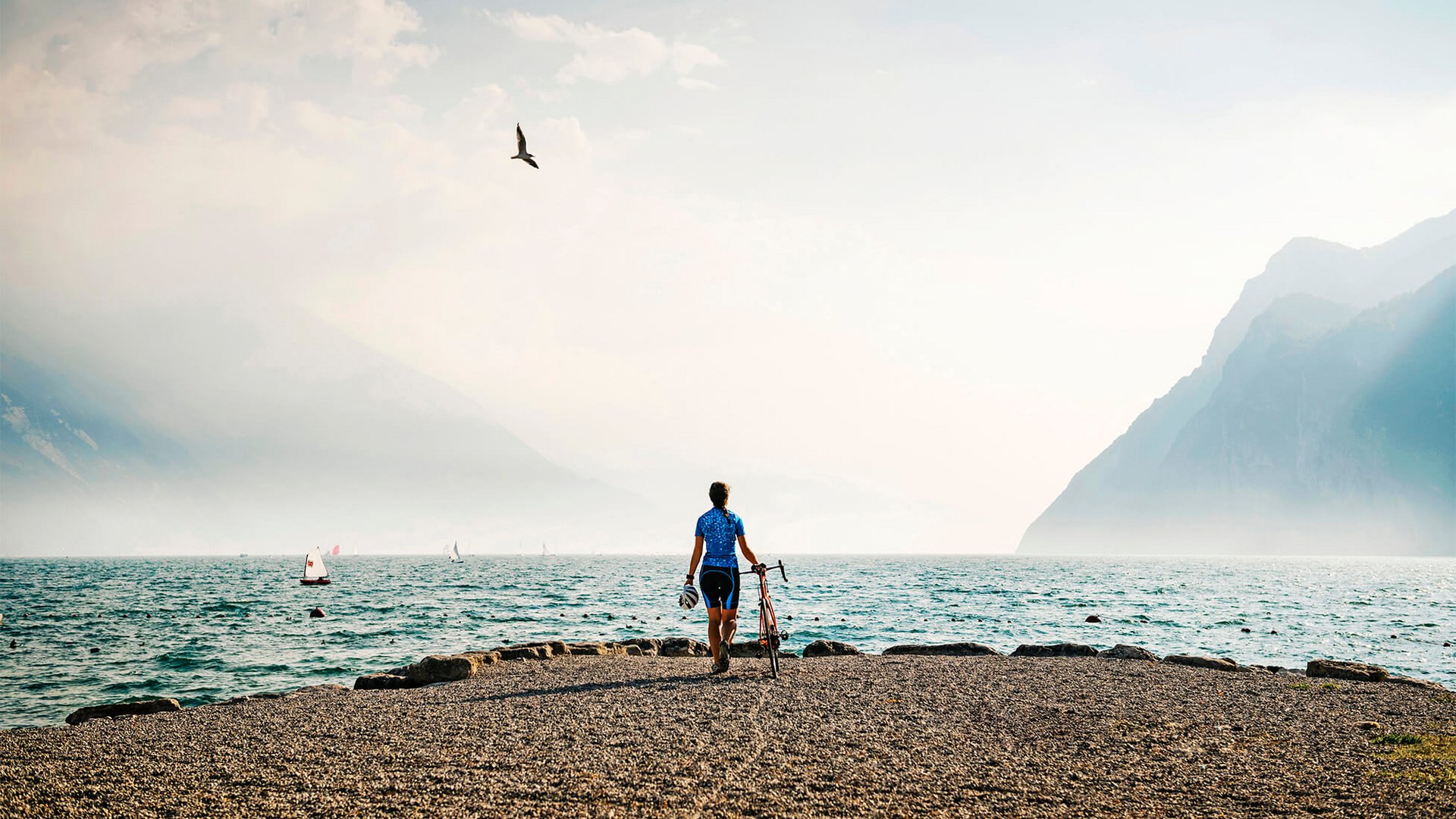 Cyclist standing by lakeside with mountains and sailboats in the background
