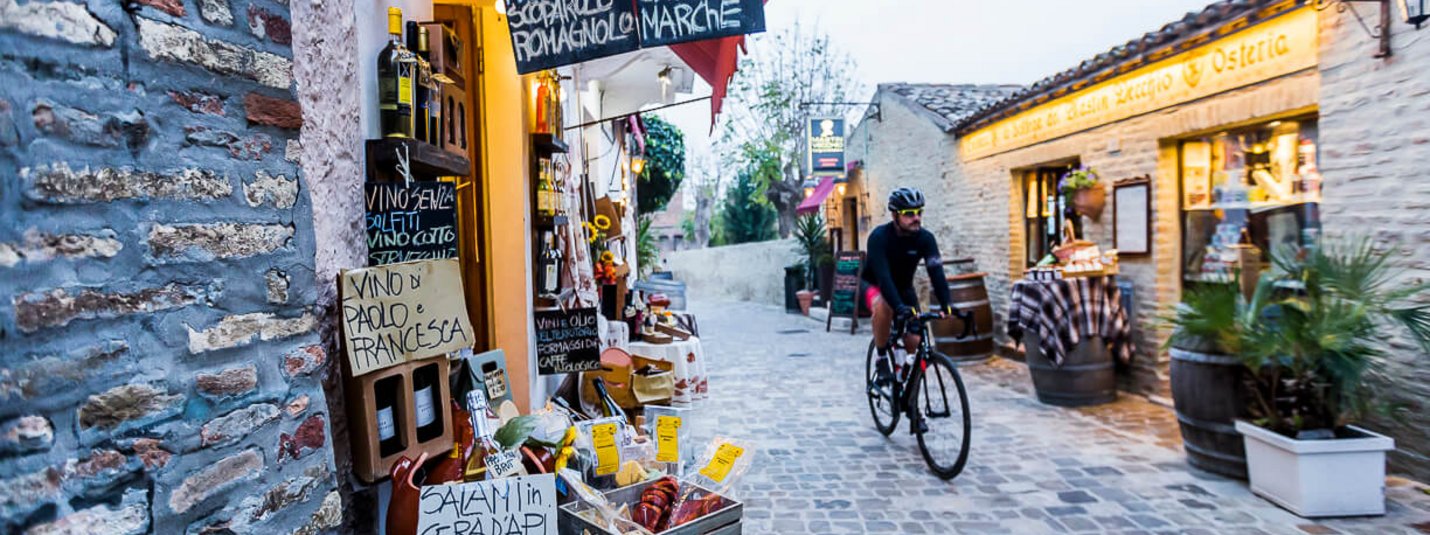 Ciclista attraversa vicolo stretto con negozio di specialità italiane