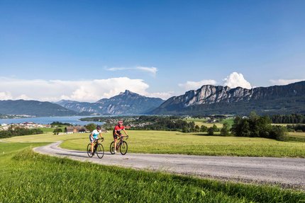 Due ciclisti su strada di campagna con montagne e lago sullo sfondo