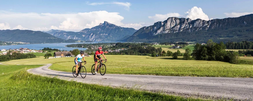 Roadbike-Bucketlist im Salzburger Land – Salzkammergut © TVB Mondsee Irrsee - Valentin Weinhaeupl Zwei Radfahrer fahren auf Landstraße mit Bergen und See im Hintergrund