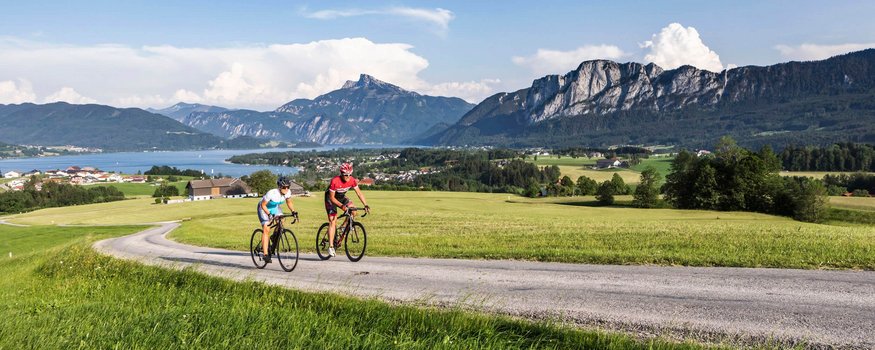 Zwei Radfahrer fahren auf Landstraße mit Bergen und See im Hintergrund