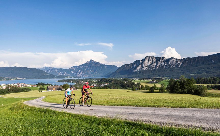 Due ciclisti su strada di campagna con montagne e lago sullo sfondo