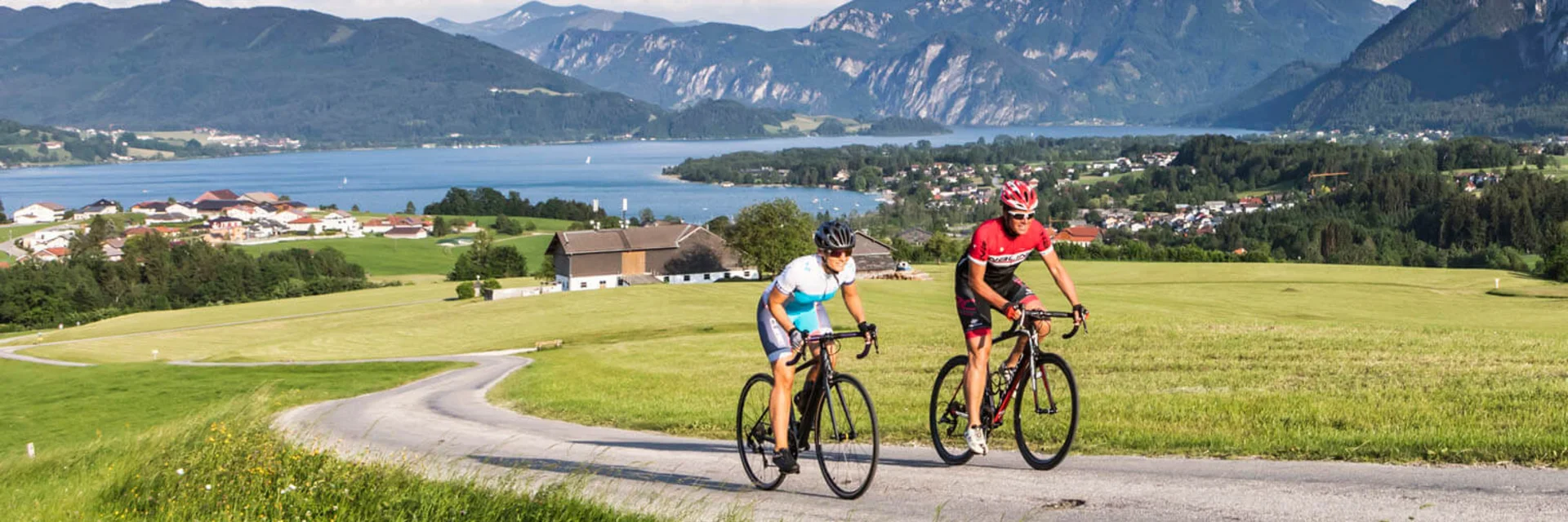 Due ciclisti su strada di campagna con montagne e lago sullo sfondo