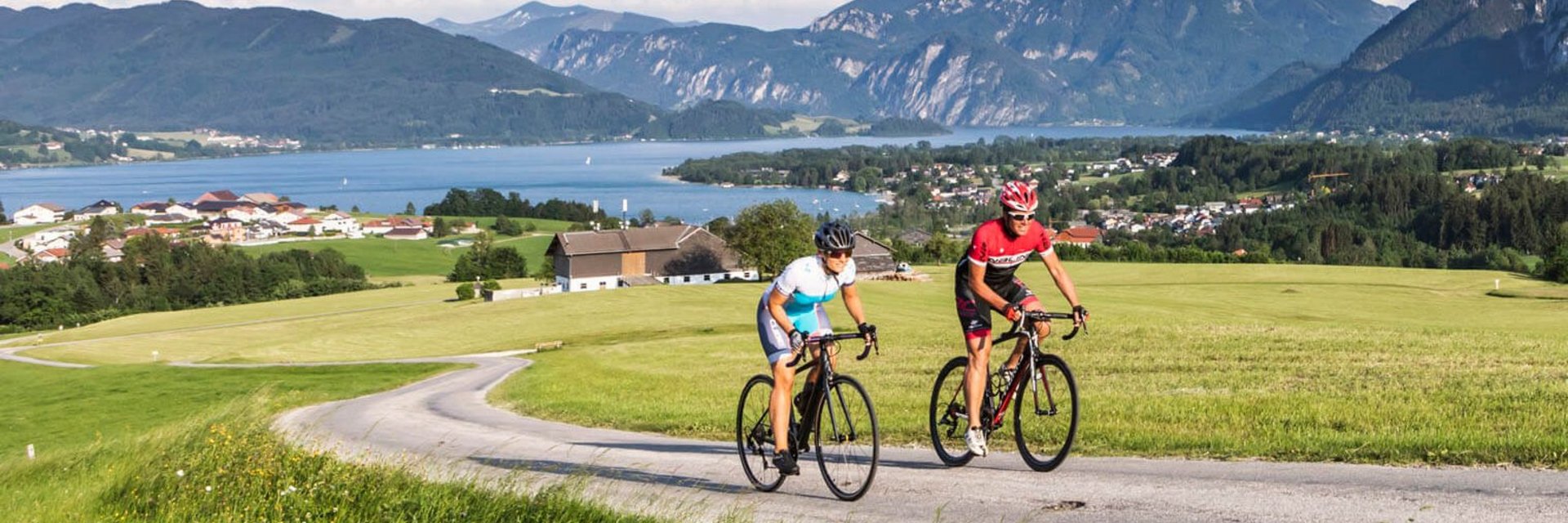 Two cyclists riding on country road with mountains and lake in background