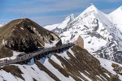 Passstraße mit Fußgängern zwischen schneebedeckten Bergen und einem Steinhaus