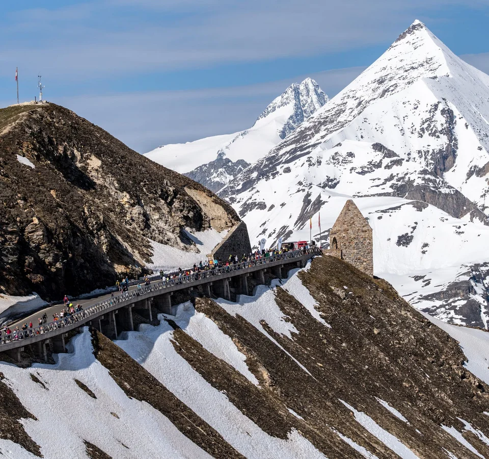 Mountain pass road with pedestrians, snowy peaks, and a stone building