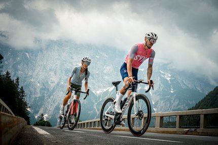 Two cyclists ride on a mountain road under cloudy sky