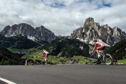 Two cyclists riding uphill in the Dolomites with village in valley
