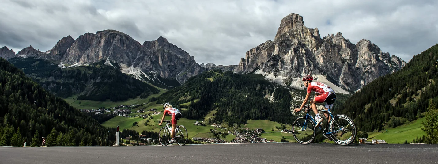 Two cyclists riding uphill in the Dolomites with village in valley