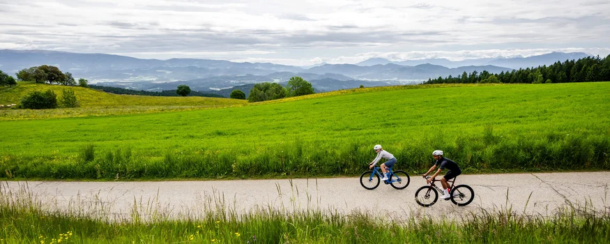 Two cyclists riding on country road with green meadows and mountains in background