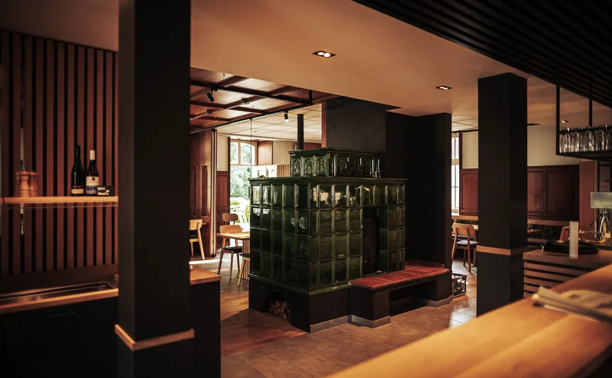 Posthorn Hotel-Ristorante Interno di ristorante con una grande stufa in maiolica verde scuro, arredi in legno e luce calda