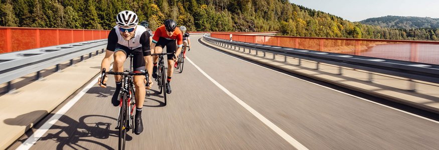 Radfahrergruppe auf Straßenbrücke mit Wald im Hintergrund bei sonnigem Wetter