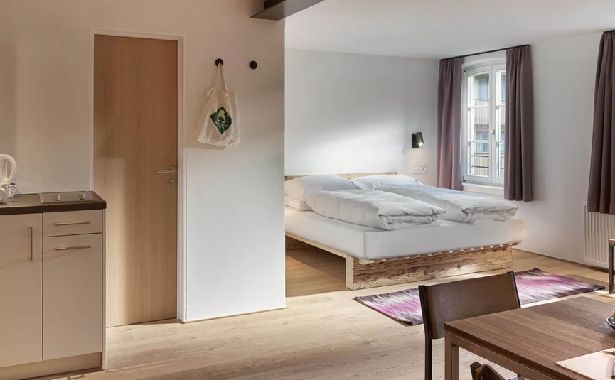 Helles Zimmer mit Küchenzeile, Esstisch und Doppelbett vor Fenstern