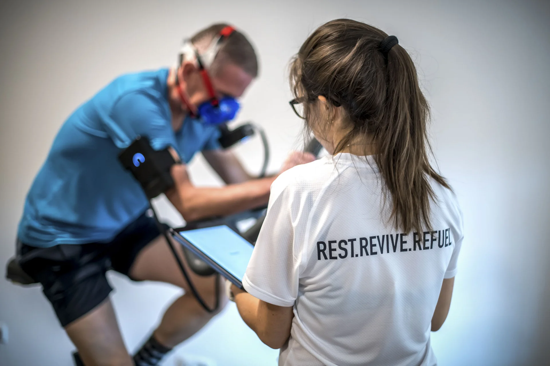 Roadbike Holidays © Fabian Schaiter Frau überwacht Mann mit Atemmaske während Training auf Fahrrad-Ergometer
