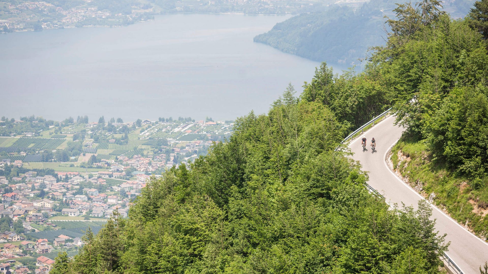 Zwei Radfahrer fahren bergauf auf kurviger Straße mit Seenblick