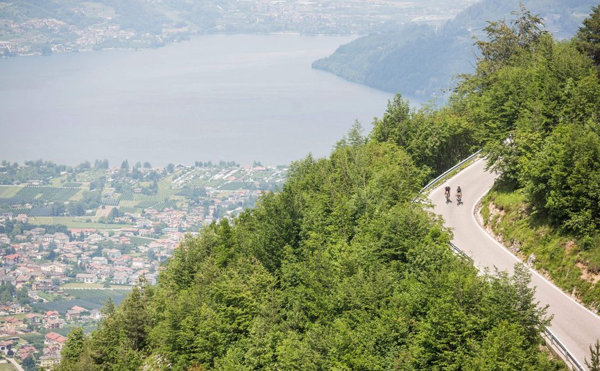 Due ciclisti in salita su strada tortuosa con vista lago