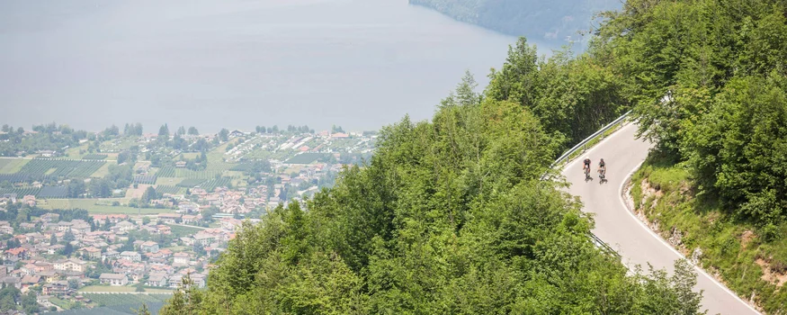 Zwei Radfahrer fahren bergauf auf kurviger Straße mit Seenblick