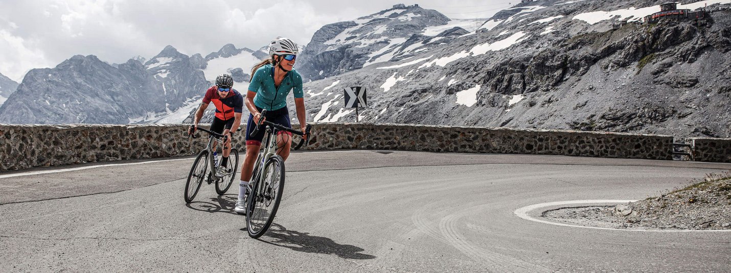 Two cyclists riding a mountain road with snow-capped peaks in the background