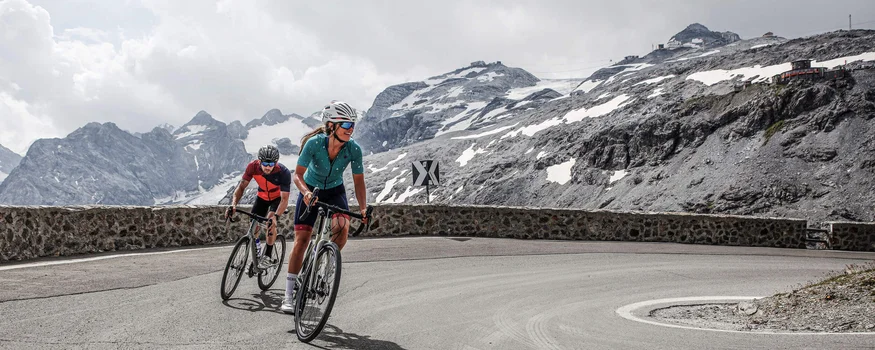 Zwei Radfahrer fahren eine Bergstraße mit schneebedeckten Gipfeln im Hintergrund