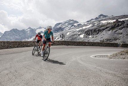 Due ciclisti su una strada di montagna con cime innevate sullo sfondo