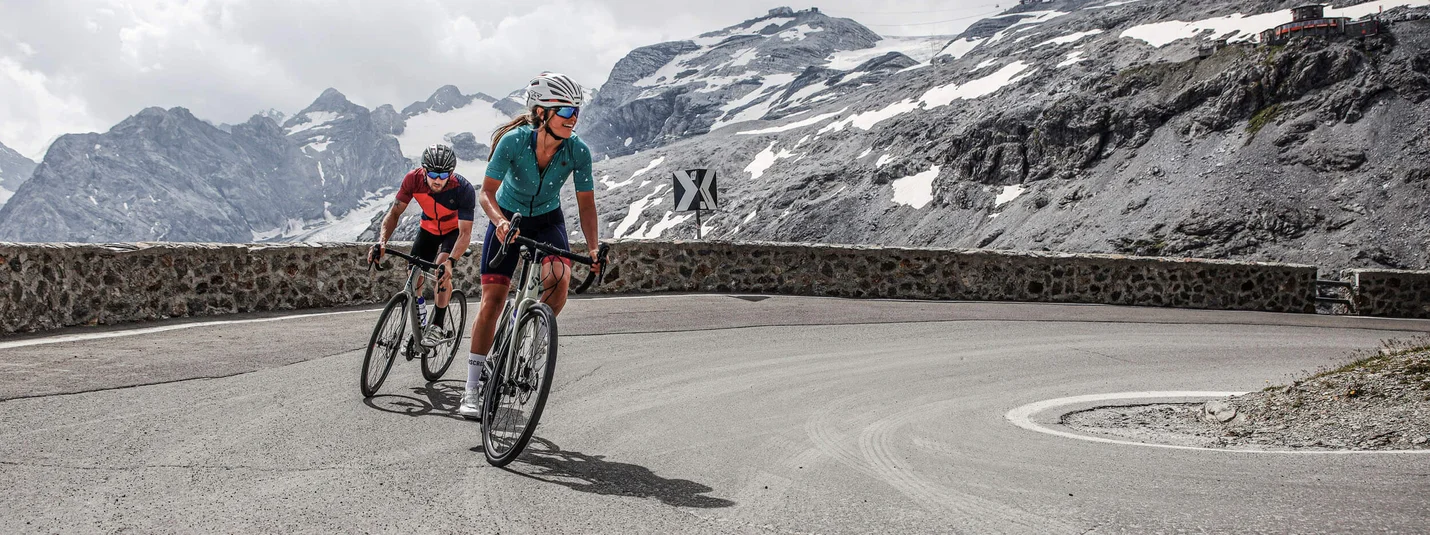 Corsa attraverso le Alpi © Felix Saller Due ciclisti su una strada di montagna con cime innevate sullo sfondo