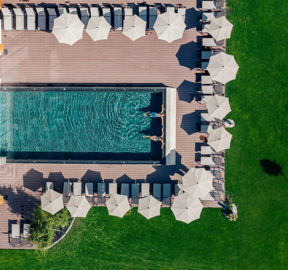 Menschen schwimmen in einem Pool mit Liegestühlen und Sonnenschirmen bei einem grünen Rasen