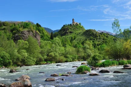 Fluss mit Steinen vor einem Hügel mit Kapelle und bewaldeten Bergen