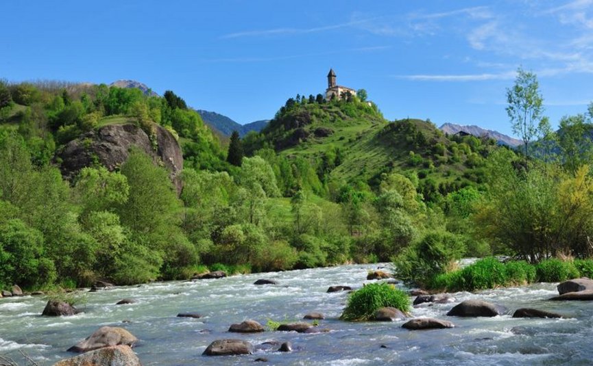 River with rocks, hill with chapel, and forested mountains in background