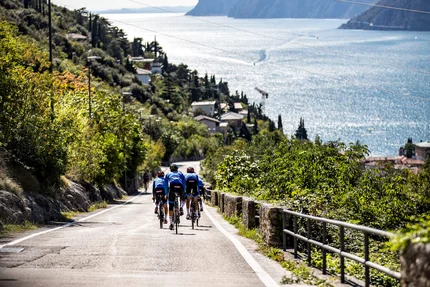 Tre ciclisti su una strada accanto a un lago con montagne sullo sfondo