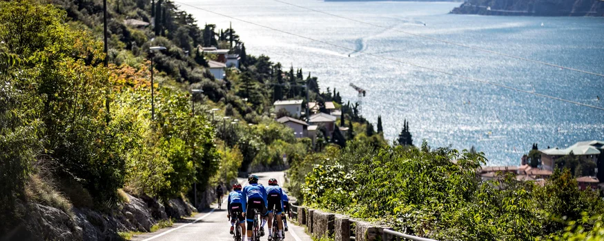 Three cyclists riding on a road by a lake with mountains in the background