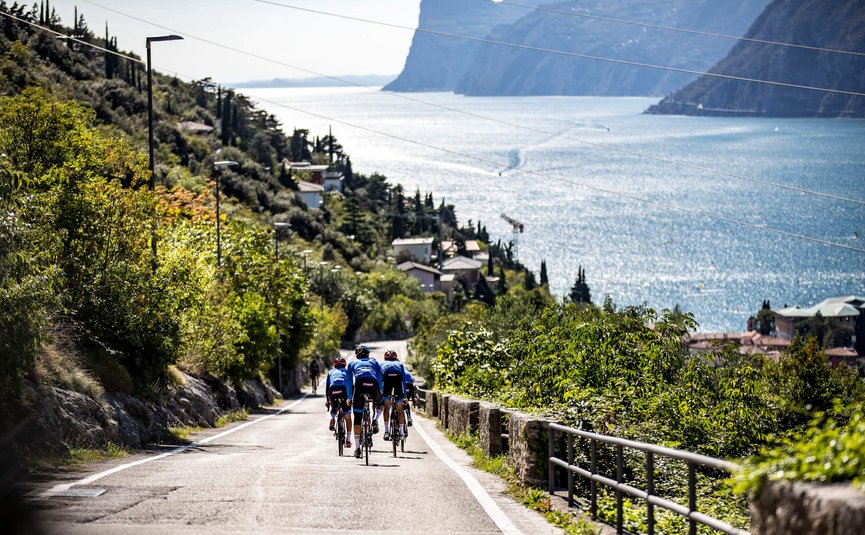 Triathlon & Rennrad Aktivhotel SantaLucia **** © Castelli Tre ciclisti su una strada accanto a un lago con montagne sullo sfondo