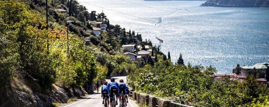 Drei Radfahrer fahren auf einer Straße entlang eines Sees mit Bergen im Hintergrund