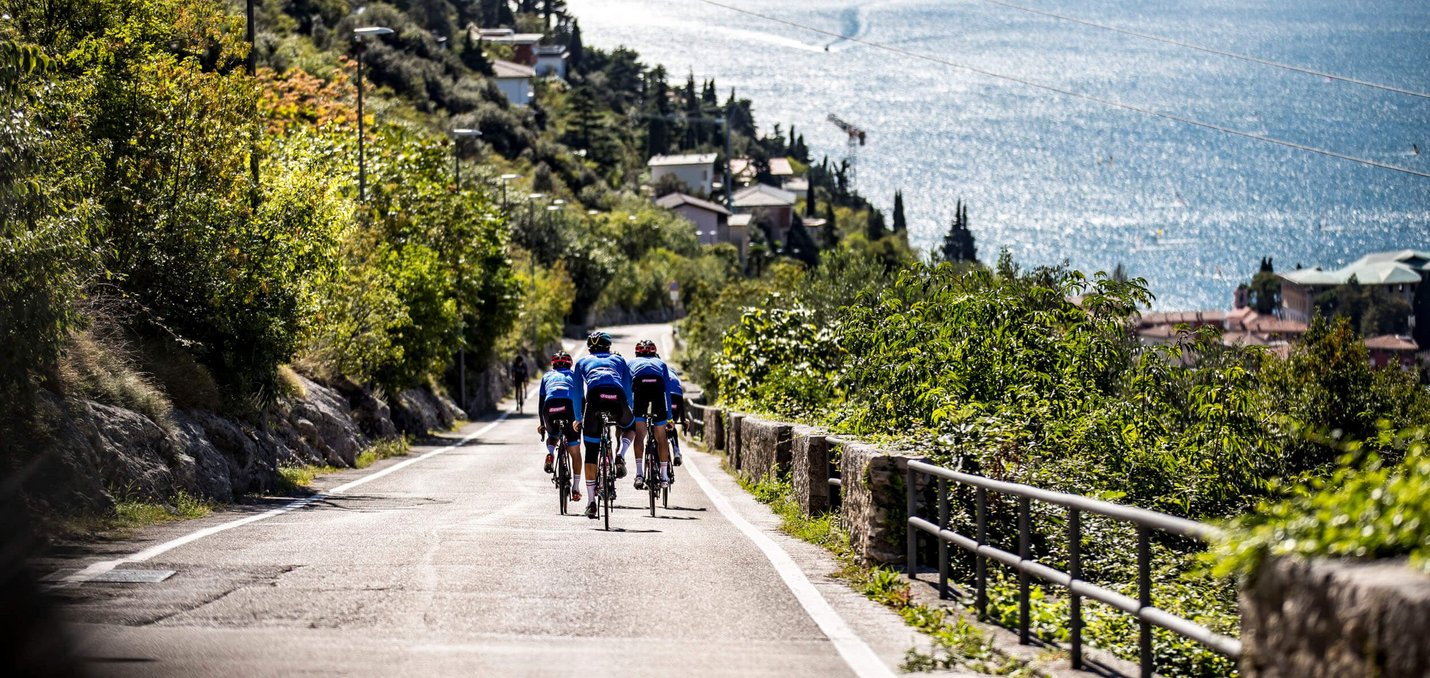 Three cyclists riding on a road by a lake with mountains in the background